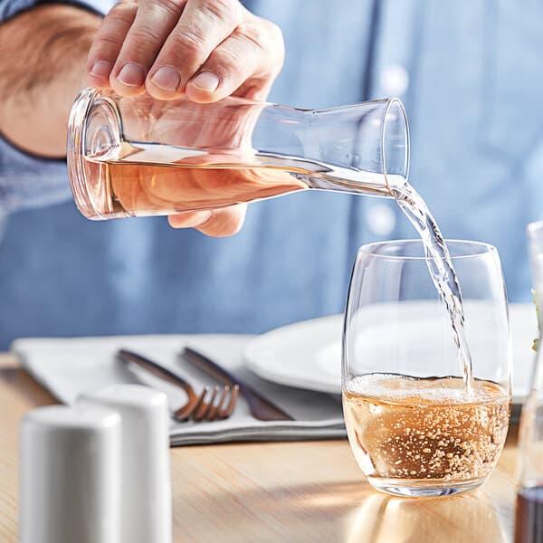 A person pouring liquid from an Acopa Slim Glass Carafe onto a table with a fork and knife.