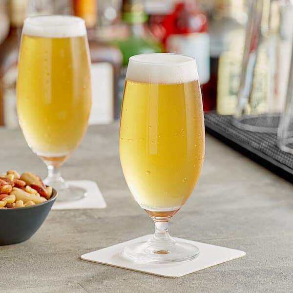 Two Acopa stemmed pilsner glasses of beer and a bowl of nuts on a table.