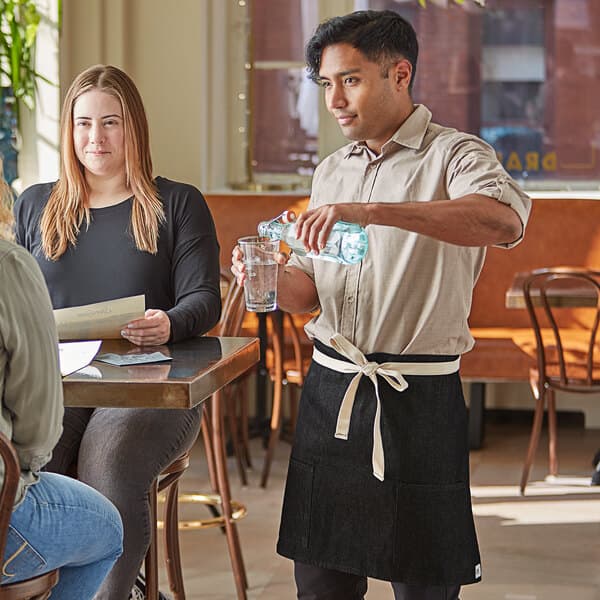 A man wearing a black denim Acopa half bistro apron pouring water at a coffee shop table.