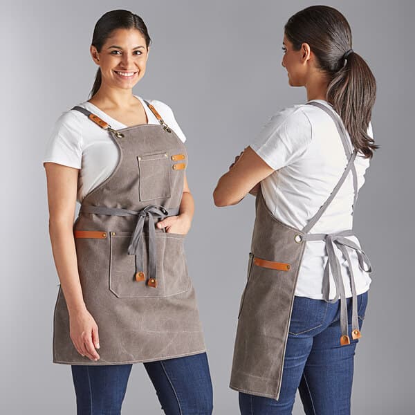 Two women wearing Acopa Hazleton canvas cross-back bib aprons in brown and another color at a counter in a professional kitchen.