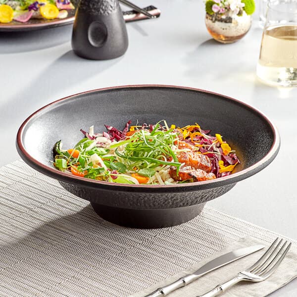 A black matte textured stoneware pasta bowl with a fork and knife on a napkin on a table.