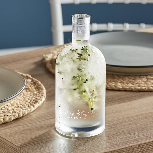 An Acopa glass bottle of liquid with ice and herbs on a table.